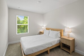 Un dormitorio con una cama, dos mesitas de noche con lámparas, una ventana con persianas y suelo alfombrado.