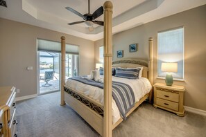 Une chambre à coucher avec un lit à baldaquin, un ventilateur de plafond, une table de chevet avec une lampe et une vue sur l’extérieur, visible à travers une porte vitrée.
