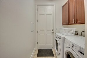 A laundry room with a white door, a washing machine, a dryer, wooden cabinets, and a laundry basket. - Fantastic 4-bedroom house with Heated Saltwater Pool & Spa (Cape Coral)