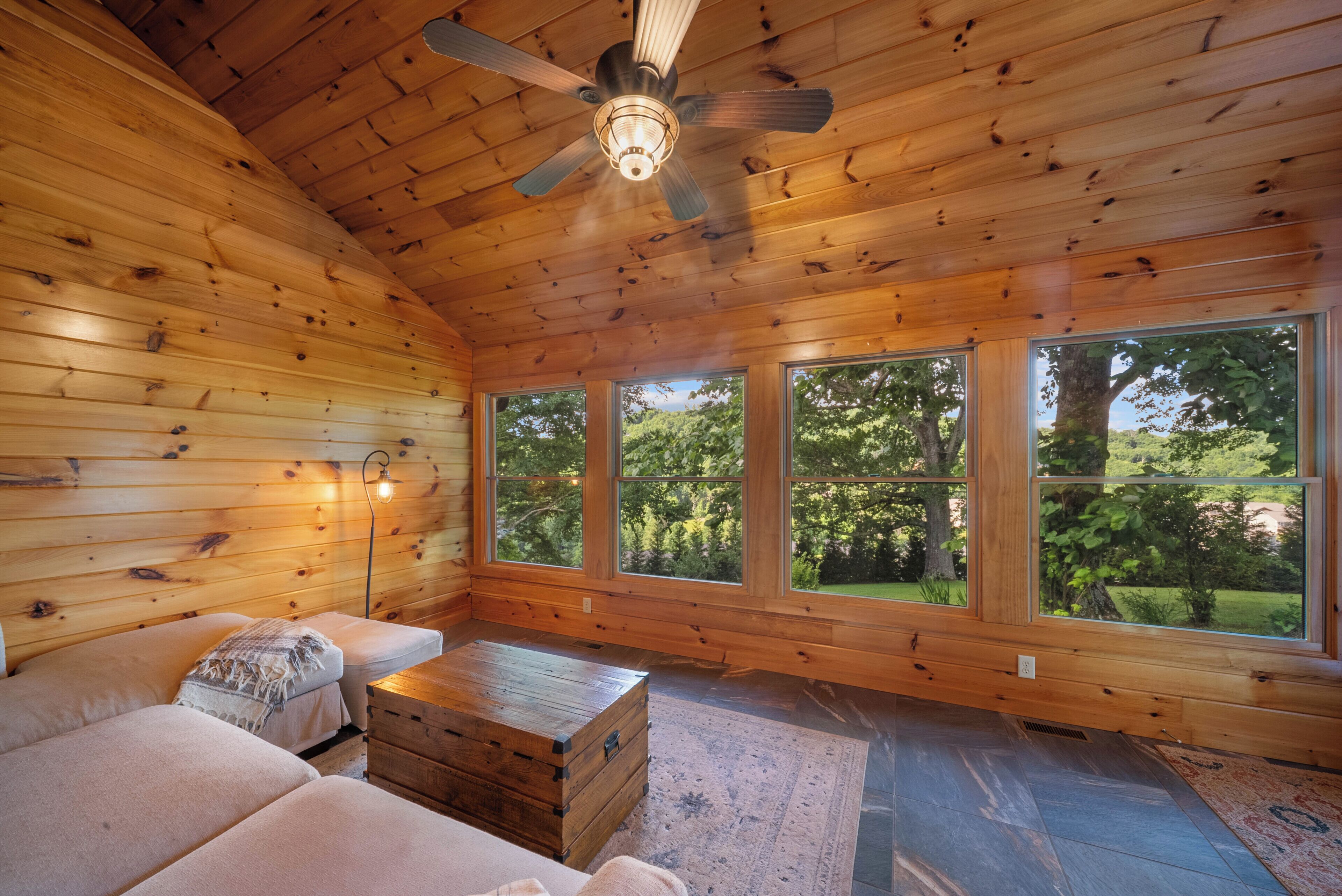 L'intérieur d'une cabane est doté d'un plafond en bois, d'un ventilateur de plafond, d'un canapé, d'une malle en bois et de grandes fenêtres offrant une vue sur la verdure.