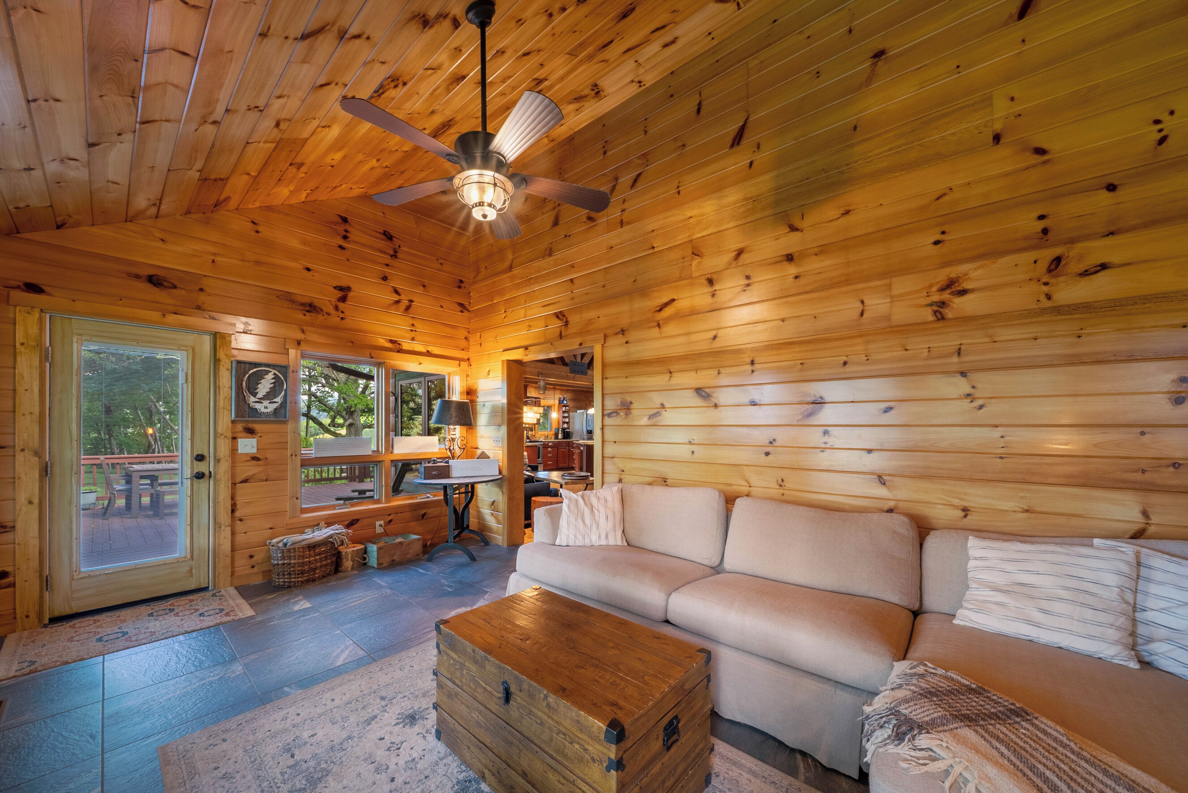 A cozy cabin interior with a wooden ceiling, a ceiling fan, a sofa, a coffee table, and a kitchen area visible through an open door.
