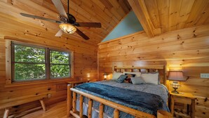 Une chambre à coucher dans une cabane en bois, avec un lit, un ventilateur de plafond, une fenêtre et une table de chevet avec une lampe.