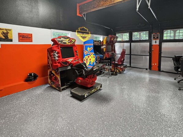 An arcade with racing game machines, a motorcycle-themed chair, and a 'Genuine Oil' sign. - Arrant Ridge - Long Range Mountain Views | Pet Friendly | Hot Tub | Firepit | Game Room (Blairsville)