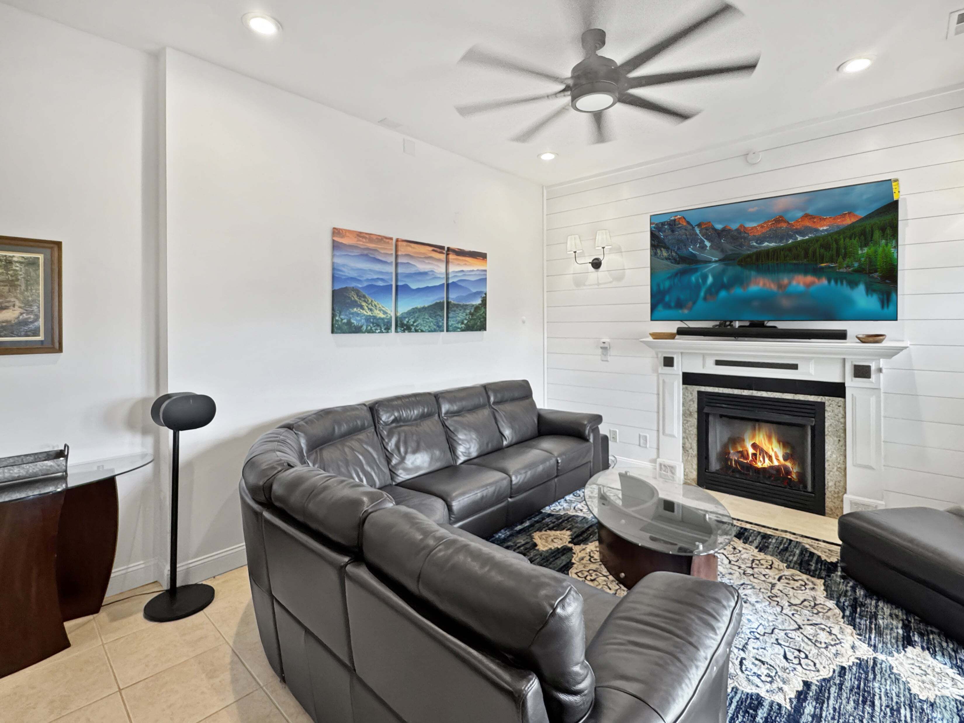 A modern living room with a leather sectional sofa, a flat-screen TV mounted above a fireplace, and a ceiling fan.