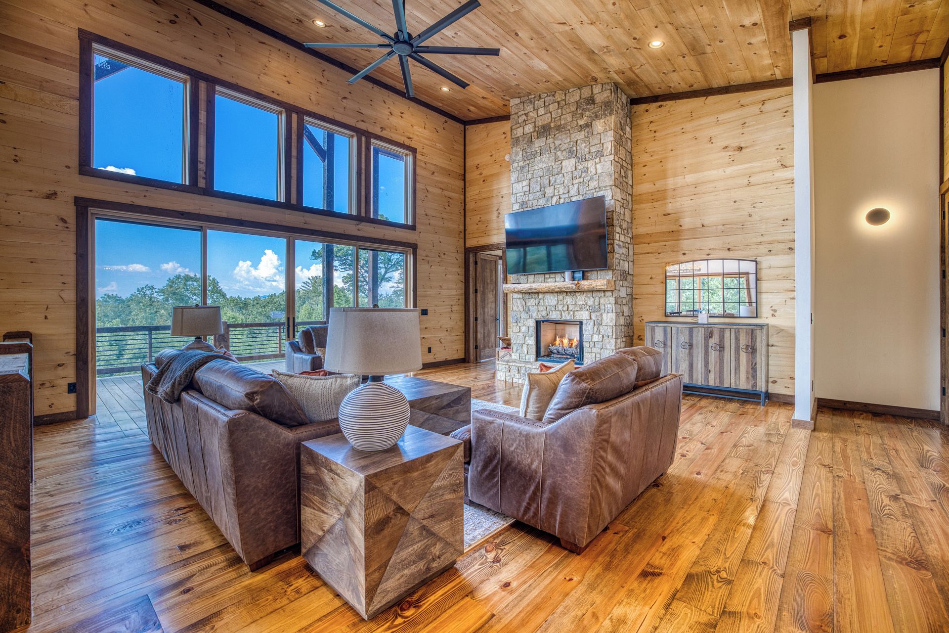 A spacious living room with a stone fireplace, leather sofas, and a large TV mounted on the wall.