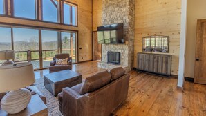 A spacious living room with a stone fireplace, leather sofas, and a large TV mounted on the wall. - Pickleball Court, Heated Pool, Mtn Views, luxury finishes, close to town (Blue Ridge)