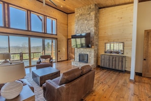 A spacious living room with a stone fireplace, leather sofas, and a large TV mounted on the wall. - Pickleball Court, Heated Pool, Mtn Views, luxury finishes, close to town (Blue Ridge)