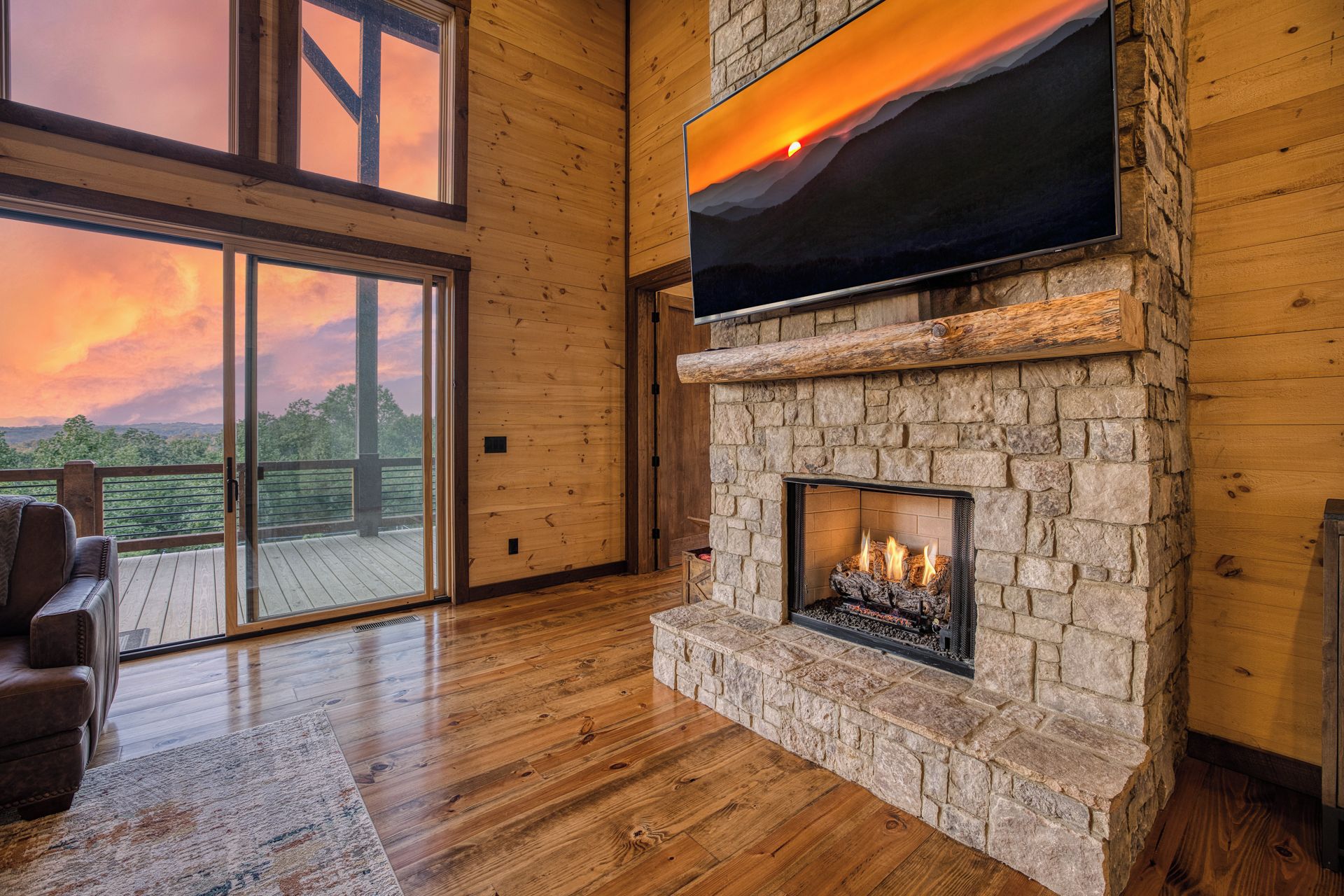 Una habitación con chimenea de piedra, un televisor grande montado encima y vista a un atardecer a través de un ventanal.