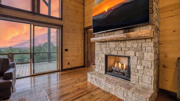 A room with a stone fireplace, a large TV mounted above it, and a view of a sunset through a glass door.