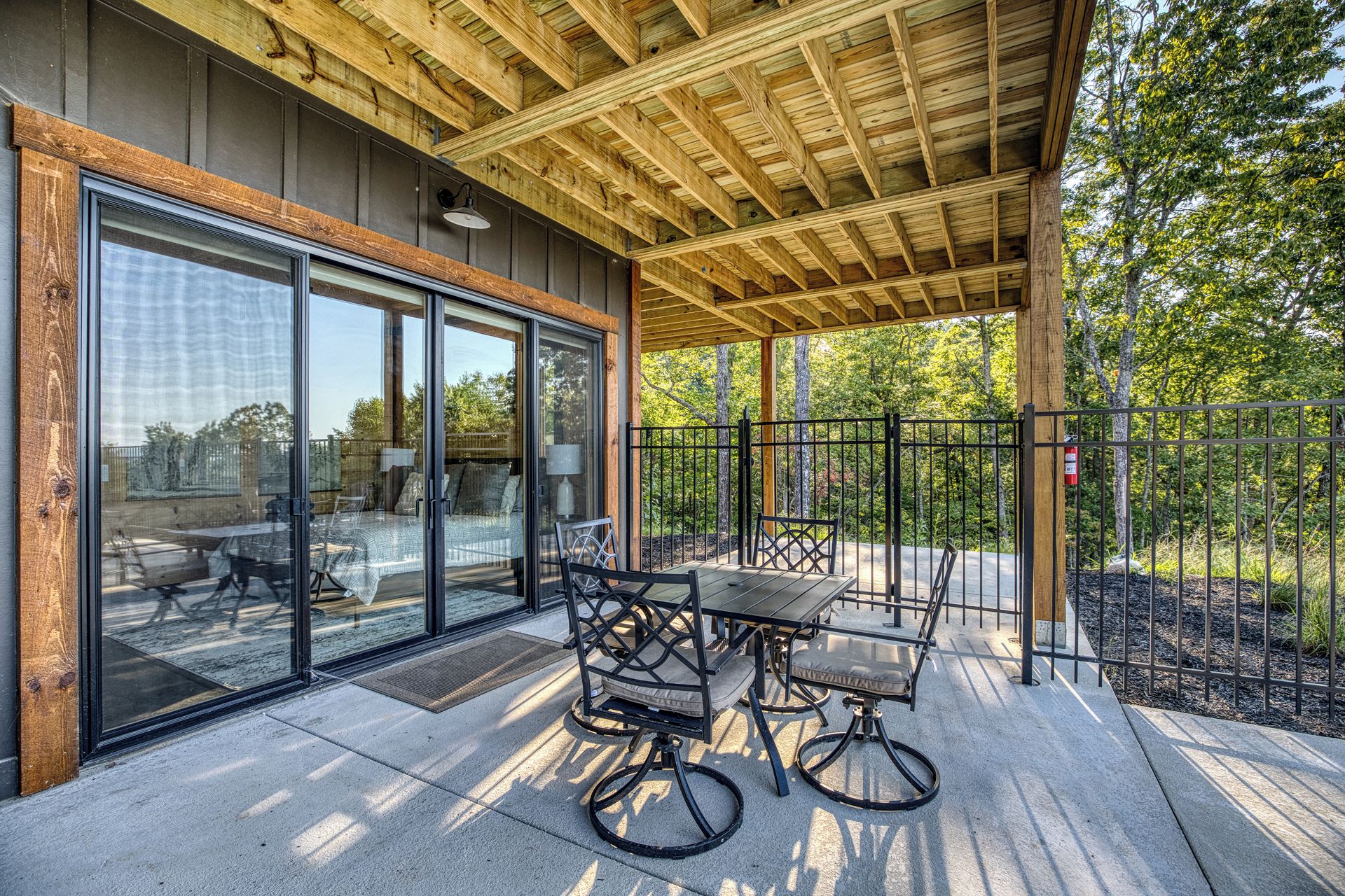 A covered outdoor seating area with a table and chairs, a glass door entrance, and a view of the surrounding trees.