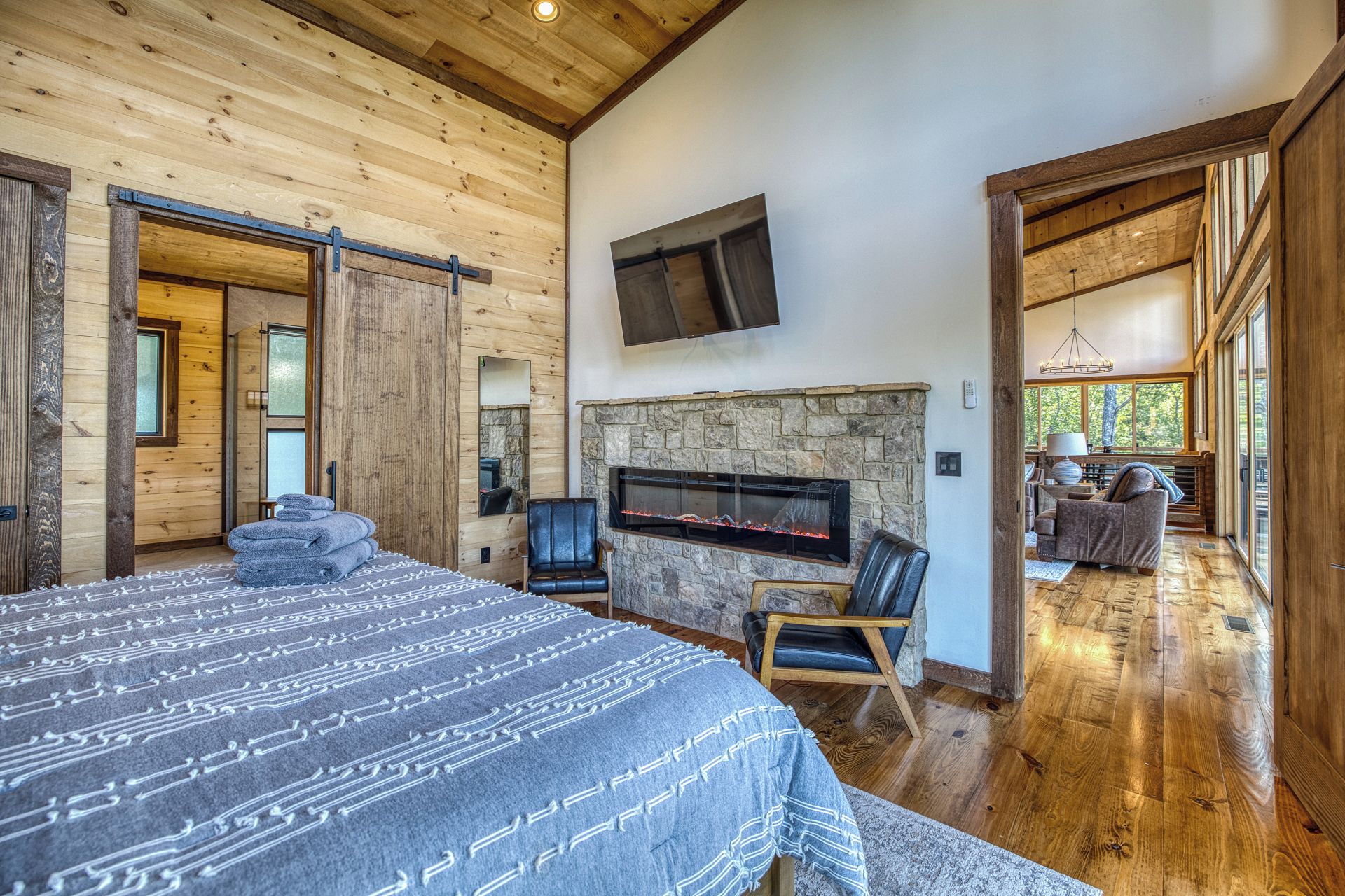 A spacious room with a large bed, a stone fireplace, a mounted TV, and a view of the outdoors.