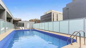 Ein Dachpool bei strahlend blauem Himmel, mit Liegestühlen und modernen Gebäuden im Hintergrund.
