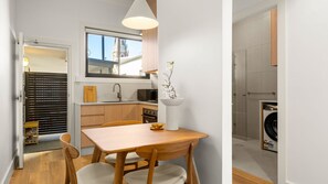 A compact kitchen with a dining area, a washing machine, and a window overlooking a building. - The Plot Twist Randwick Unit 2 (Randwick)
