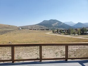 Blick vom Balkon mit Holzwertung auf eine ländliche Landschaft, mit einem Berg im Hintergrund und einer kleinen Siedlung mit Häusern und einer Straße.