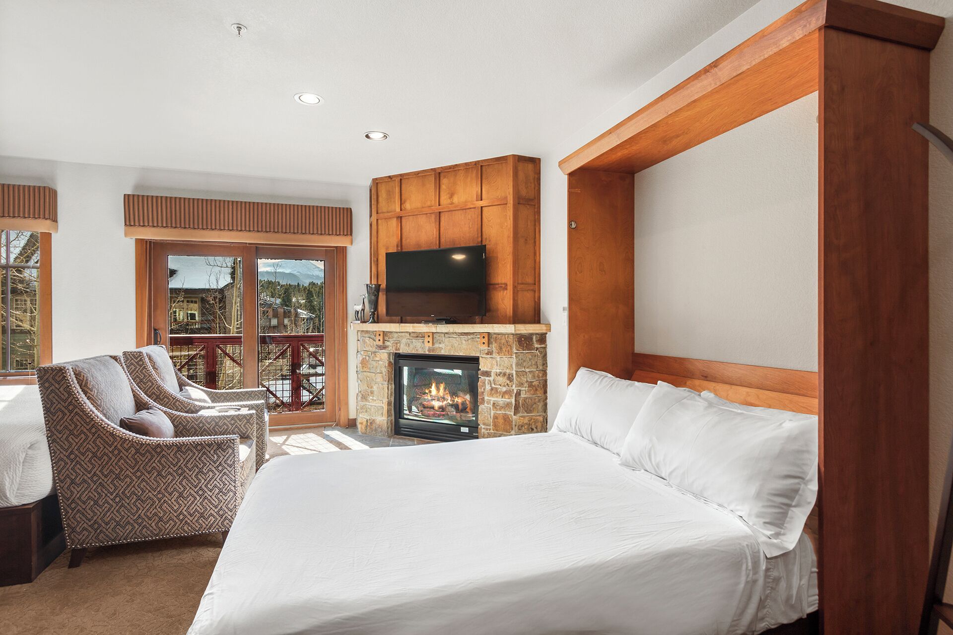 A bedroom with a large bed, a fireplace, a TV, and a balcony with chairs.