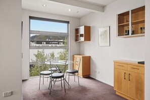 A modern room with a glass table and chairs, a wooden cabinet, and a large window offering a view of a building and trees. - Penthouse Apartment in the heart of Nelson (Nelson)