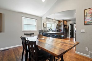 A dining area with a wooden table and chairs, a kitchen in the background, and a window with blinds. - Spacious Golden Home Near Red Rocks & DT with EV (Golden)