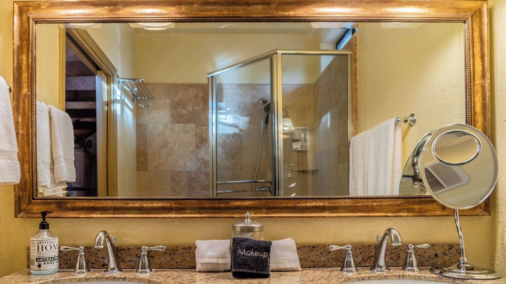 Un bagno con uno specchio grande, un lavandino con due rubinetti, una bottiglia di disinfettante per le mani e un barattolo con l'etichetta 'Makeup'.