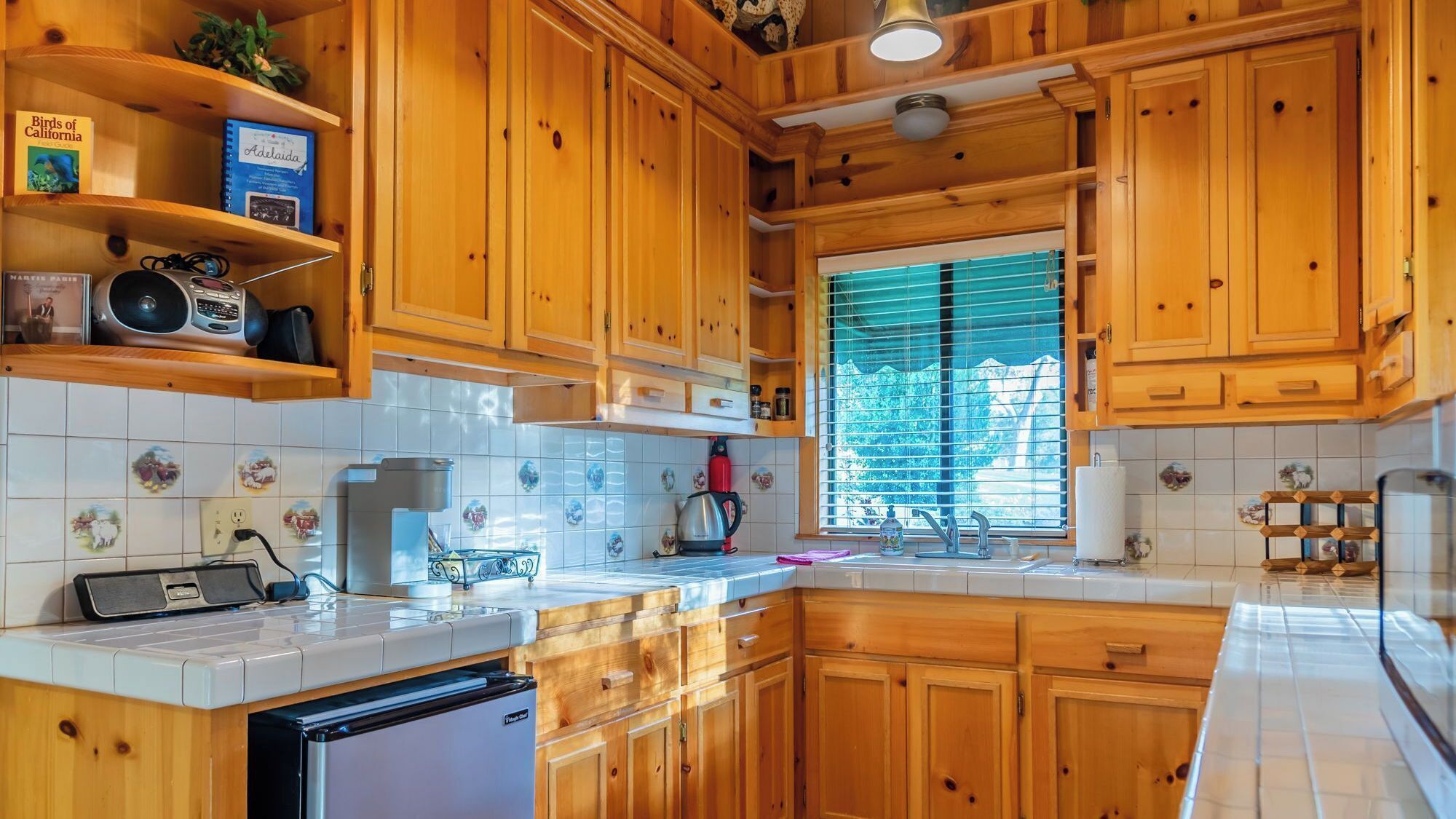 A kitchen with wooden cabinets, a built-in oven, a coffee maker, and a window with blinds.