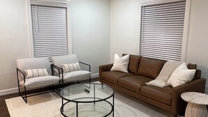 Una sala de estar con un sofá marrón, dos sillones, una mesa de centro de vidrio y una mesita auxiliar.