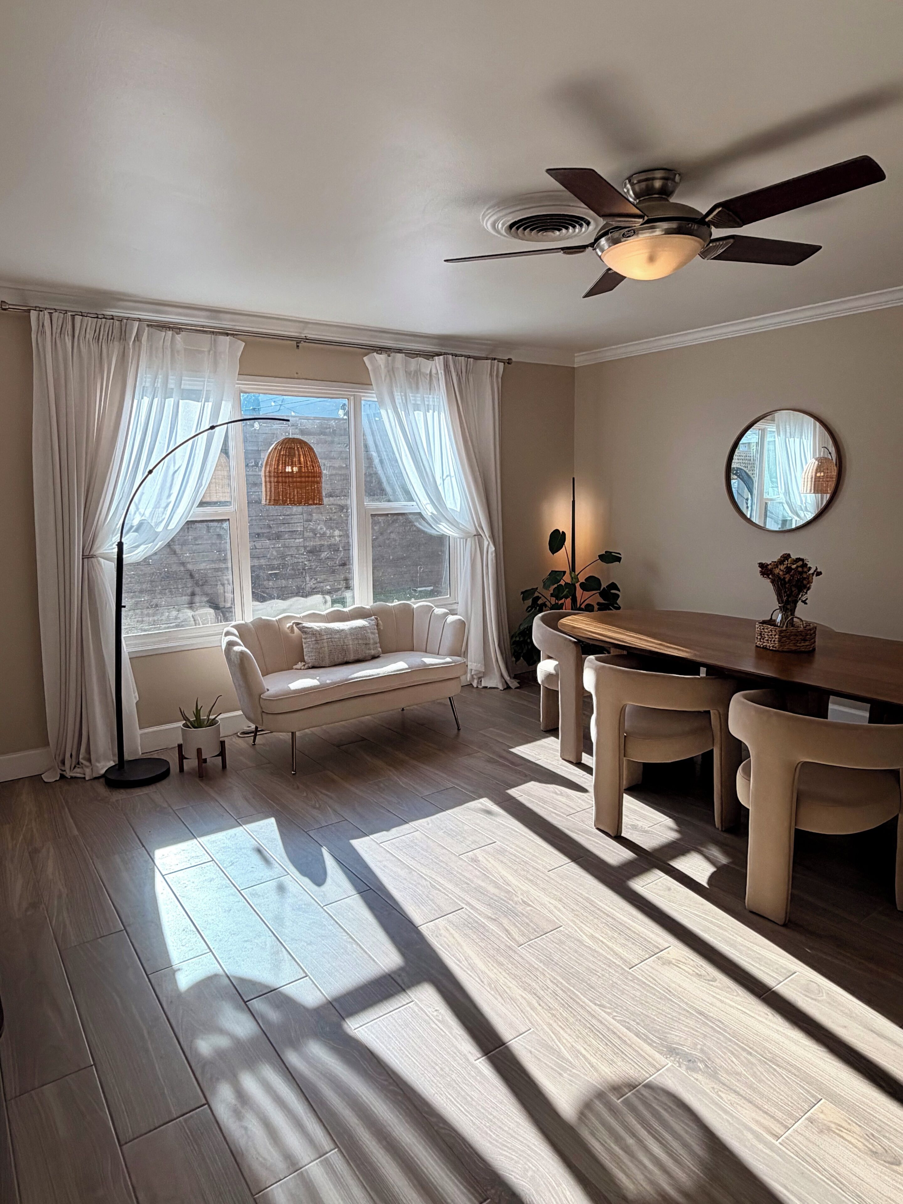 A room with a ceiling fan, a sofa, a dining table with chairs, a window with curtains, and a potted plant.