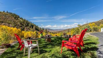 An outdoor seating area with a table and chairs, surrounded by a grassy lawn and a view of a forested hillside.