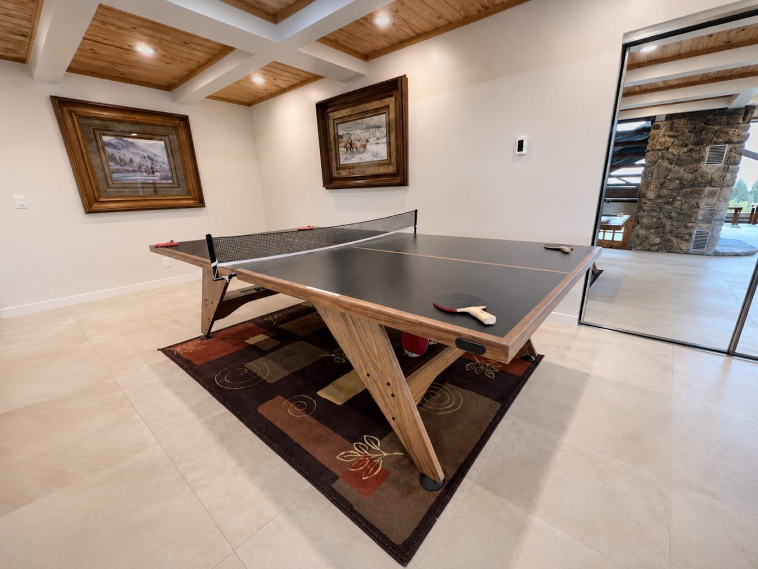 Une table de ping-pong dans une pièce dont le plafond est orné de poutres en bois, les murs sont décorés de tableaux encadrés et un pilier en pierre se dresse dans un coin.