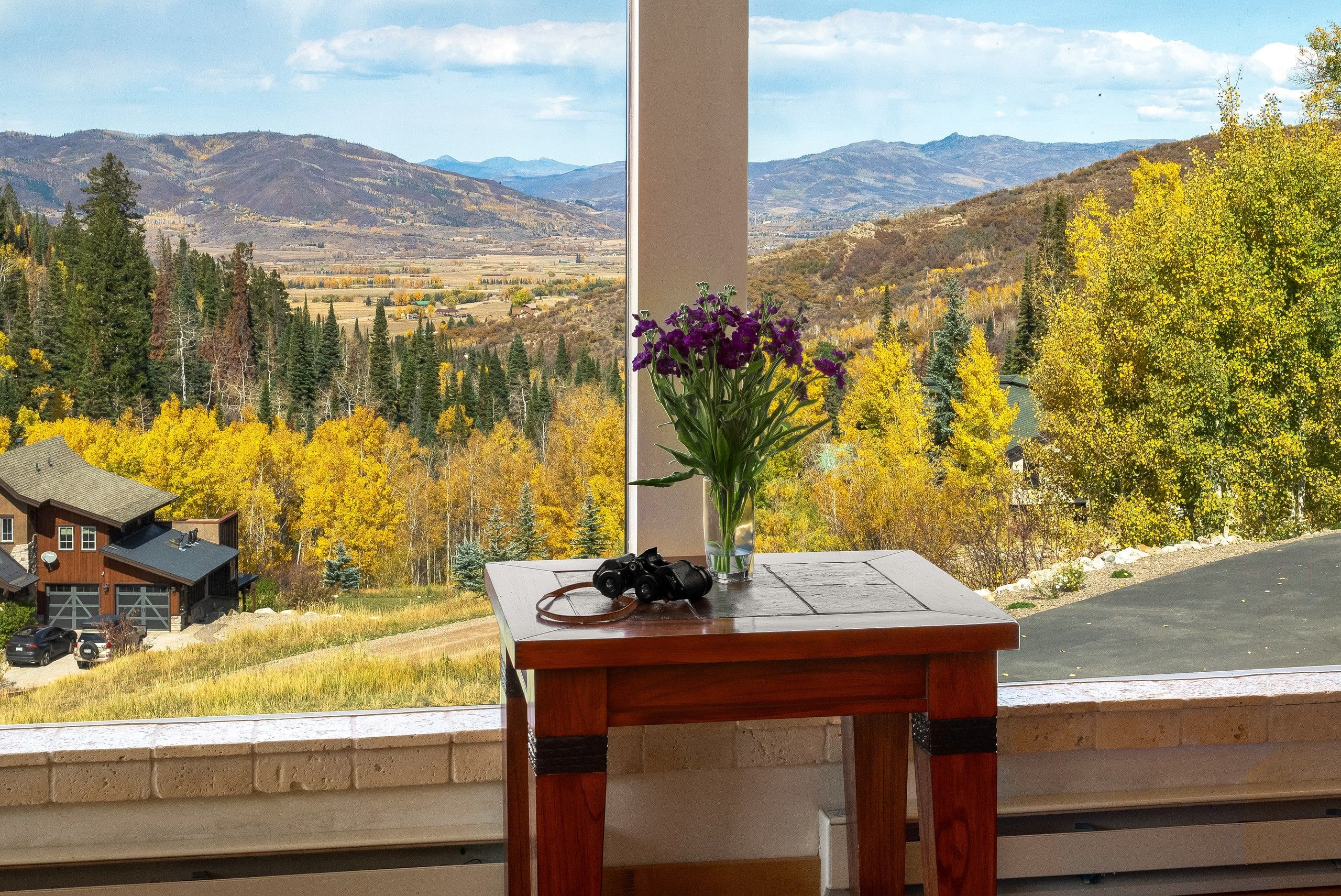 Una mesa de madera con una cámara y flores, con vistas a un paisaje montañoso con follaje otoñal.