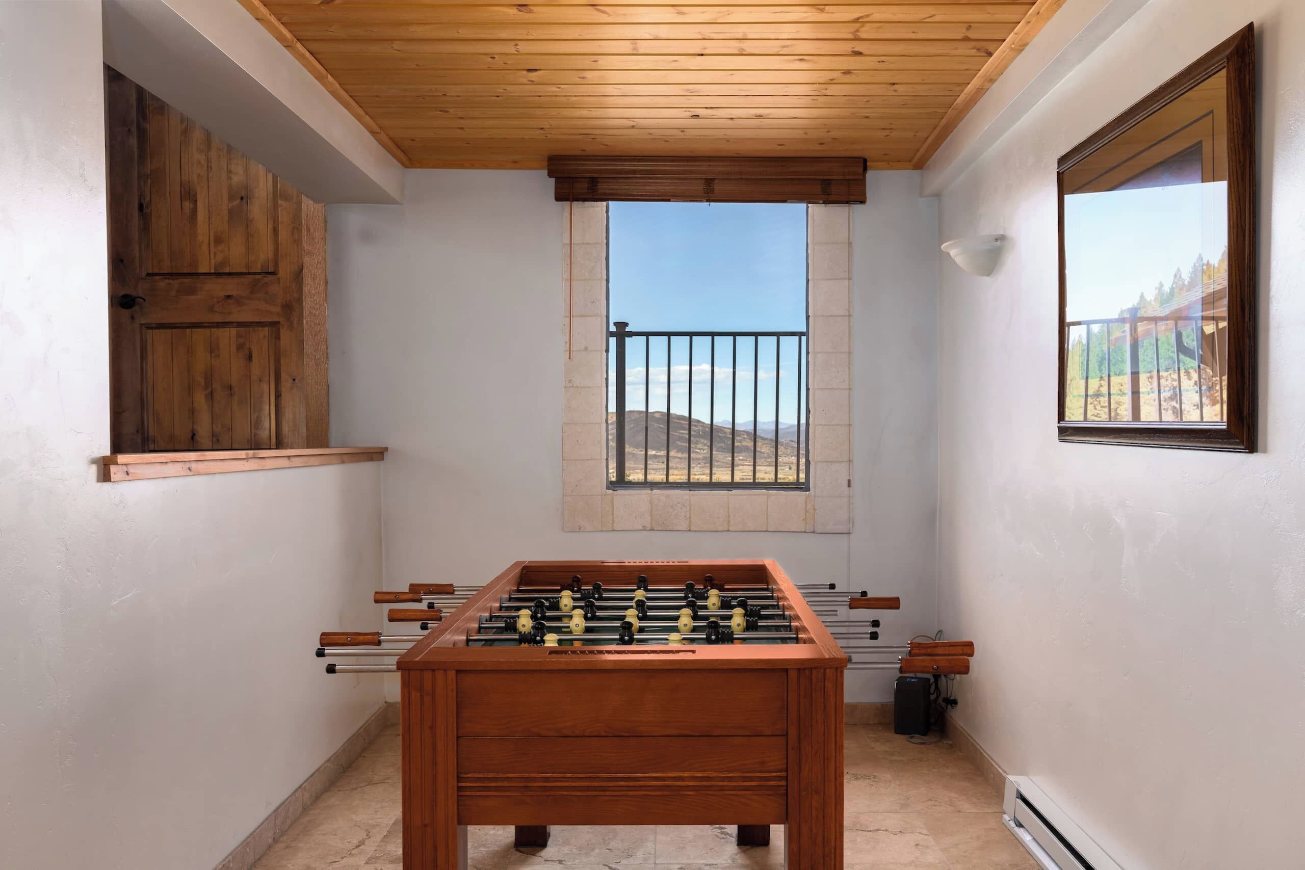 Una habitación con mesa de fútbol, ventana con vistas a colinas y puerta de madera.