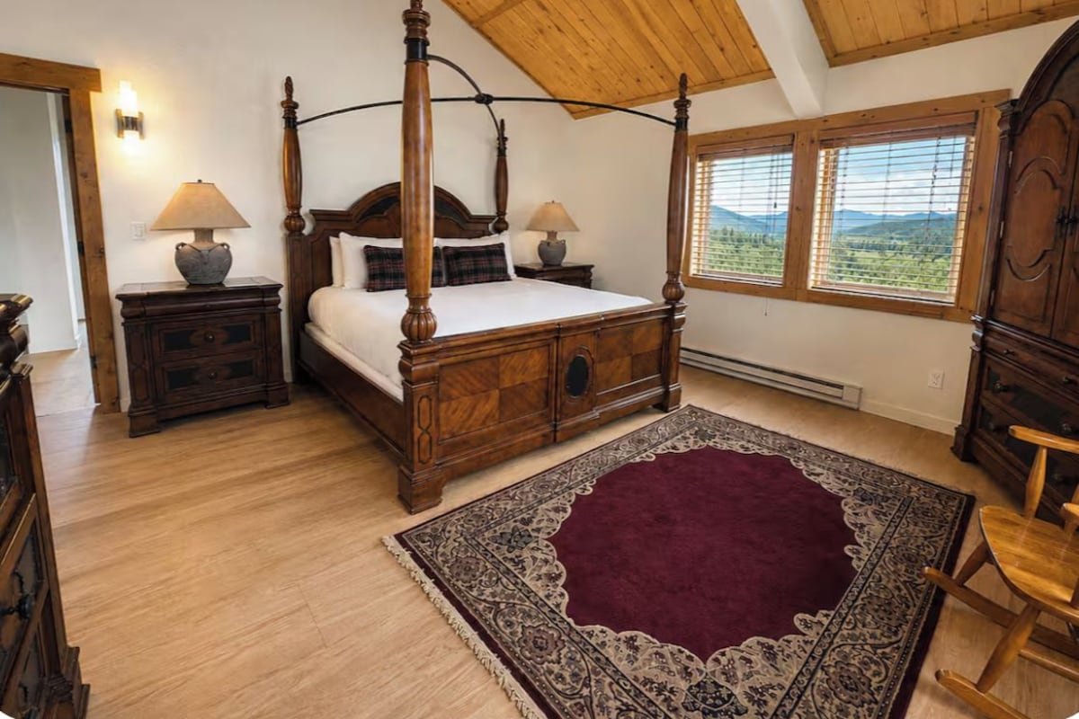 A four-poster bed with a wooden headboard, two bedside tables with lamps, a large patterned rug, and a view of the outdoors through a window.