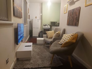 Olohuoneessa on litteänäyttöinen televisio, kuviollisia koristetyynyjä sisältävä sohva, pieni valkoinen sivupöytä ja taustalla näkyy keittiö.