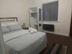 A hotel room with a bed, a bedside table, a lamp, a hanging coat rack, a window with curtains, and a radiator.