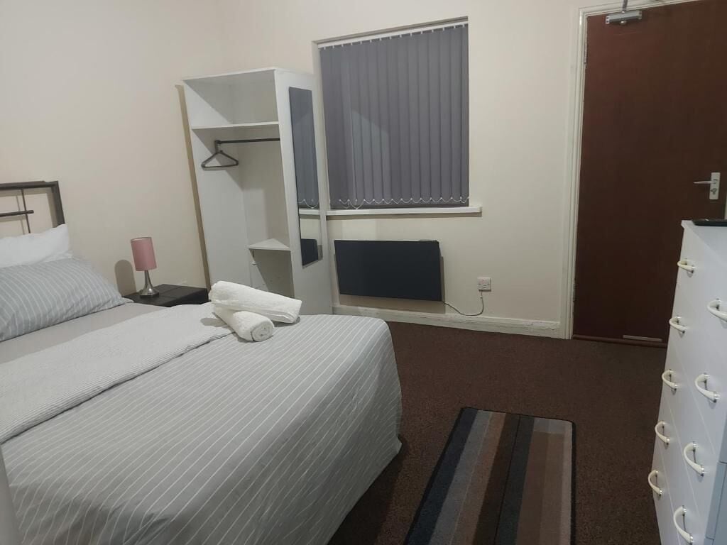 Habitación de hotel con cama, mesita de noche, espejo, ventana con cortinas, radiador y armario.