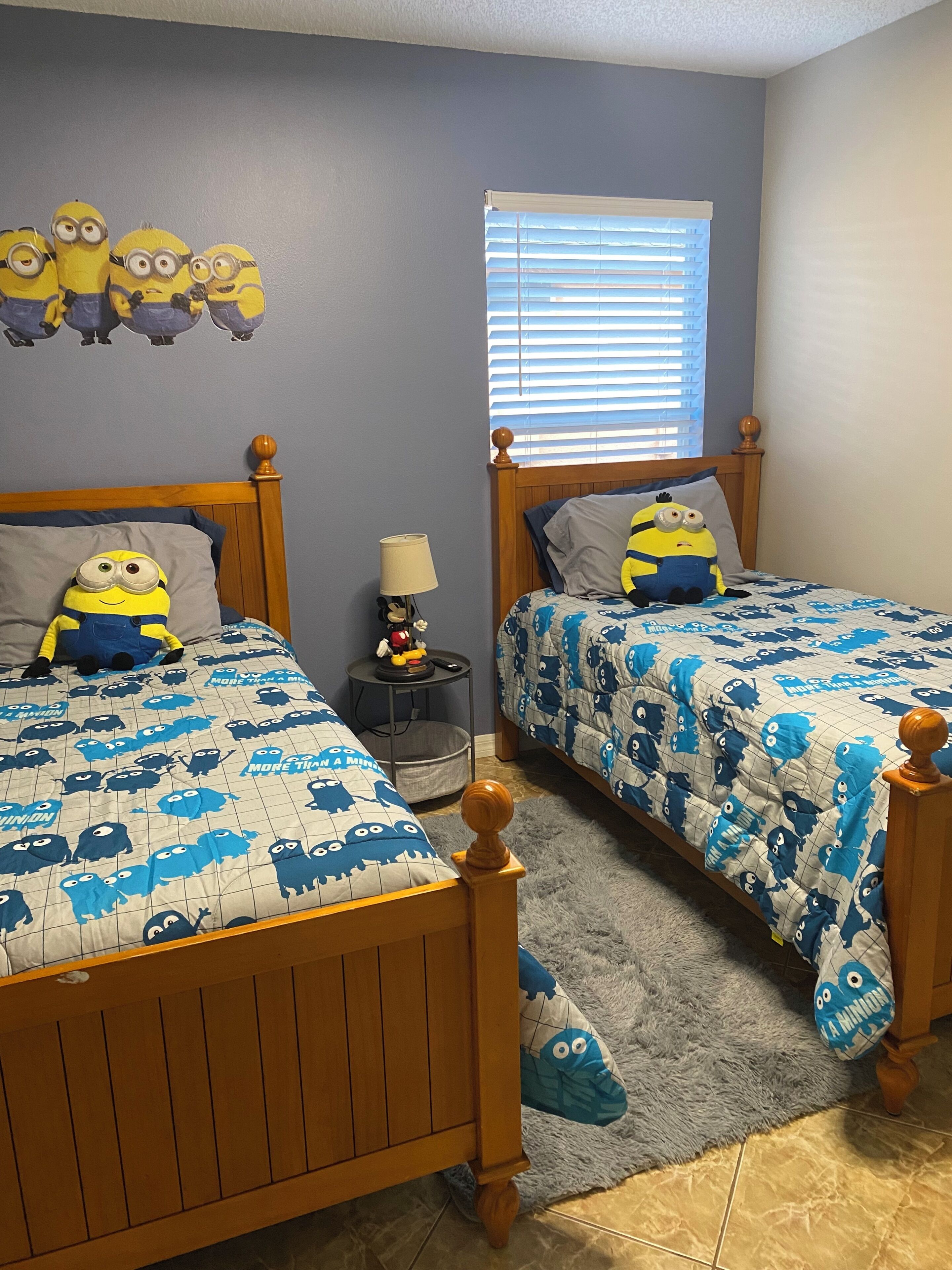 A bedroom with two beds, each with a Minion-themed bedspread, a nightstand with a lamp, and a window with blinds.