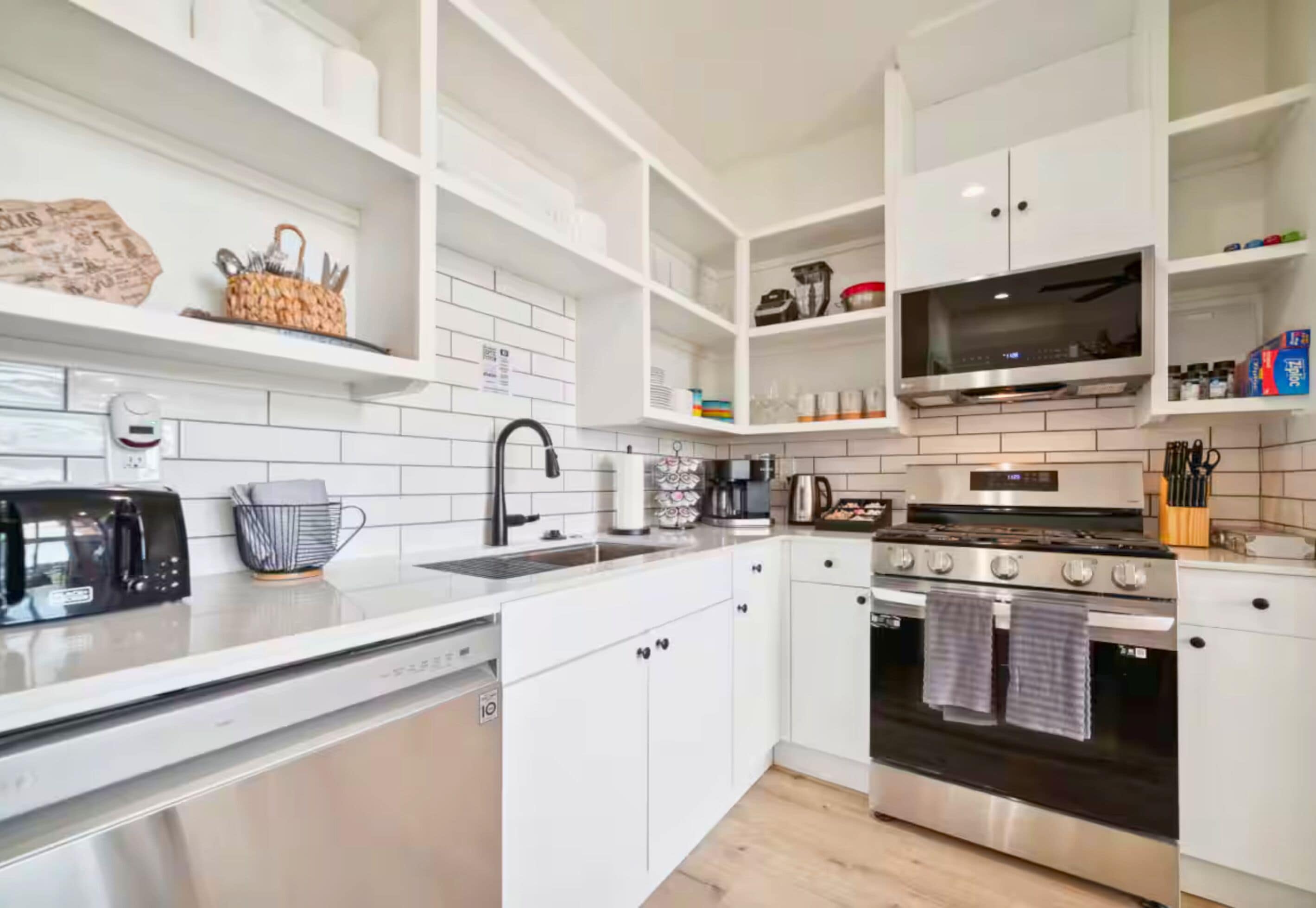 A modern kitchen with white cabinetry, a stainless steel dishwasher, a black microwave, a coffee maker, a kettle, and a stove.