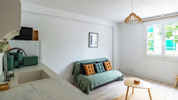 Une pièce avec un canapé vert, une petite table ronde, un luminaire suspendu, un tableau encadré au mur et une fenêtre donnant sur un paysage verdoyant.