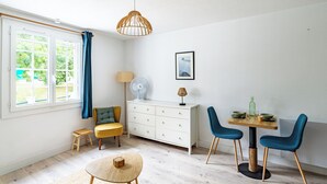 Une pièce comprenant une table à manger, des chaises, un petit tabouret, un meuble, un ventilateur, une lampe et une fenêtre avec des rideaux.