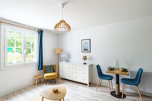 A room with a dining table, chairs, a small stool, a cabinet, a fan, a lamp, and a window with curtains. - Studio Baignol - Welkeys (Ciboure)
