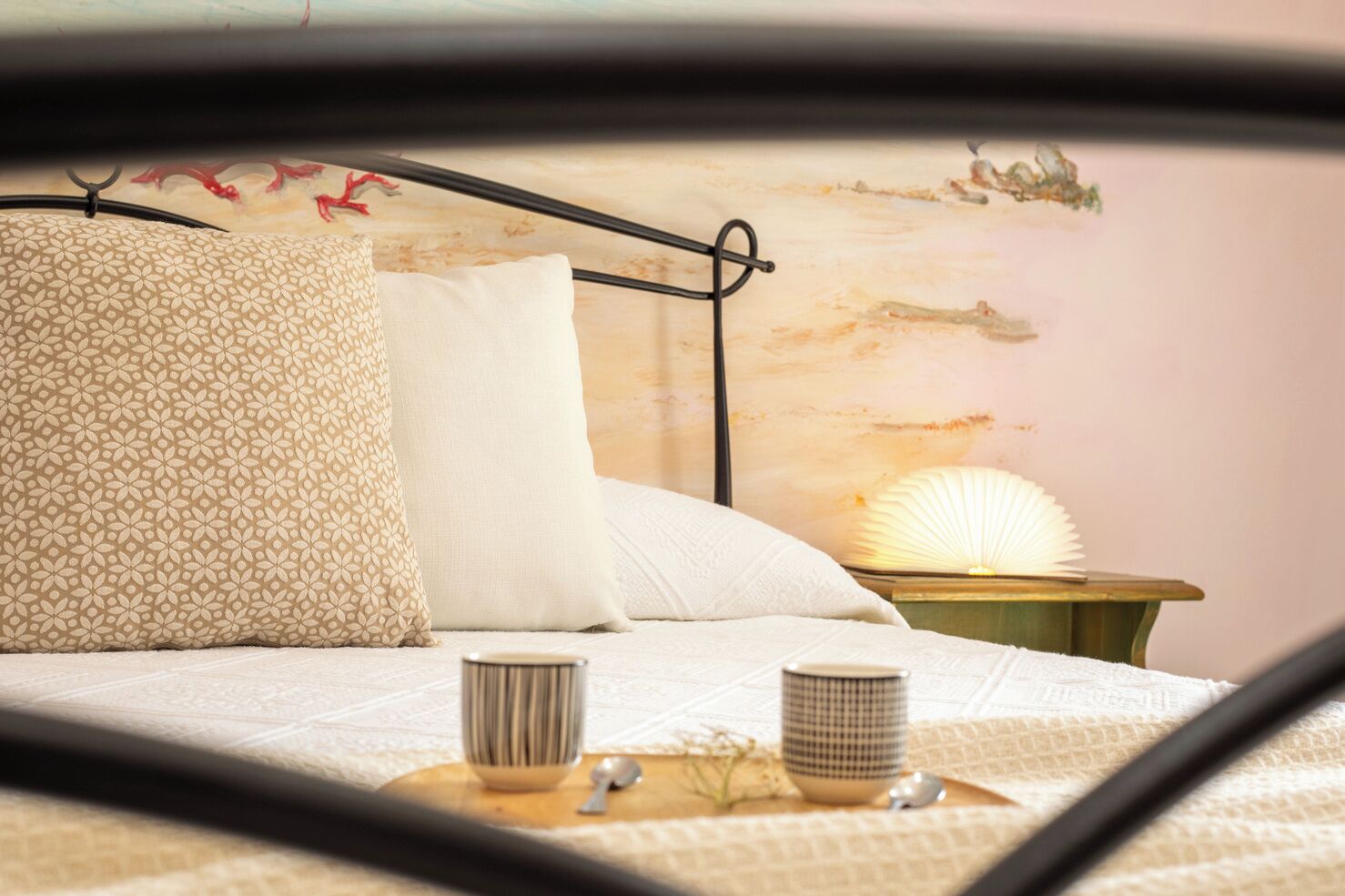 A bed with a patterned pillow, a plain pillow, and a bedside table with a lamp and two cups.