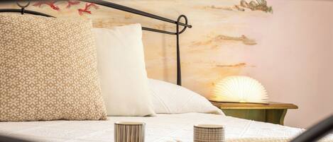 A bed with a patterned pillow, a plain pillow, and a bedside table with a lamp and two cups.