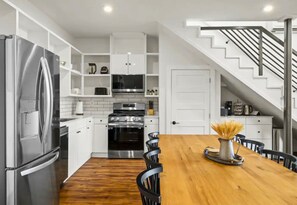 Eine moderne Küche mit weißen Schränken, Edelstahlgeräten, einem Holzespeise Tisch und einer Treppe, die in einen oberen Bereich führt.