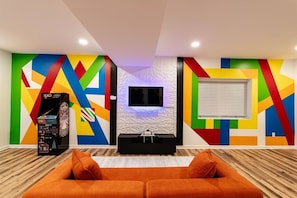 A modern living room with a colorful geometric wall design, a flat-screen TV, and a wooden floor. - 5BR Mass Resort | Movie Lounge +Pool/Poker Table+Arcade+Gym+Remote Workstatio... (McGaheysville)