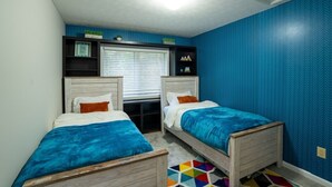 Quarto com duas camas, uma janela com persianas, uma estante com objetos decorativos e uma parede estampada em azul.