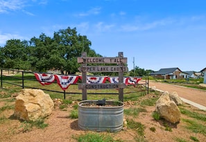 A wooden signpost with 'WELCOME Y'ALL' and 'UPPER LAKE CABINS' written on it, surrounded by a metal barrel and flags. - A-Frame Cabin | Community Ponds | Fire Pit (New Braunfels)