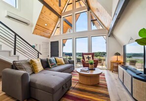 A spacious living room with a high ceiling, a sofa set, a coffee table, and a large window offering a view of the outdoors. - 3 Side by Side A-Frame Cabins | Sleeps 24 | Ponds (New Braunfels)