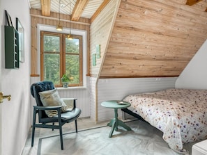 Une chambre avec un plafond en pente, un lit recouvert d’une literie à motifs floraux, une chaise avec un coussin, une petite table et une fenêtre donnant sur un paysage verdoyant.