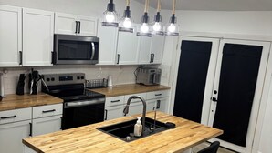 A modern kitchen with white cabinets, a wooden island with a sink, and a black faucet. There are two black bar stools and a stainless steel microwave above the island.