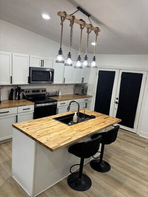 A modern kitchen with white cabinets, a wooden island with a sink, and a black faucet. There are two black bar stools and a stainless steel microwave above the island.