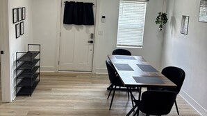 A room with a wooden floor, a dining table with chairs, a black metal shelf, a door with a curtain, and a window with blinds.