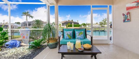 A spacious balcony with a view of a swimming pool, a table with drinks and snacks, and a teal sofa with decorative pillows.